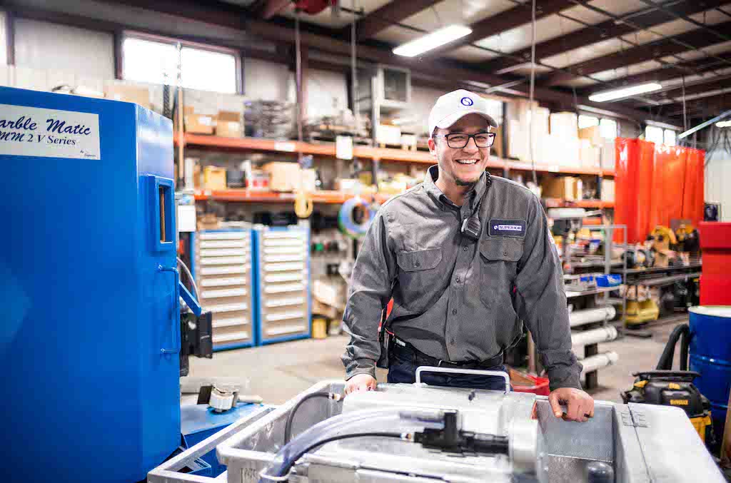 Smiling technician in safety glasses and Superior uniform operating machinery in an industrial chemical workspace.