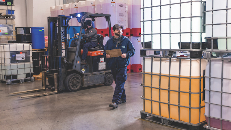 Chemical warehouse worker with clipboard overseeing forklift operations and chemical totes storage for inventory management and logistics.